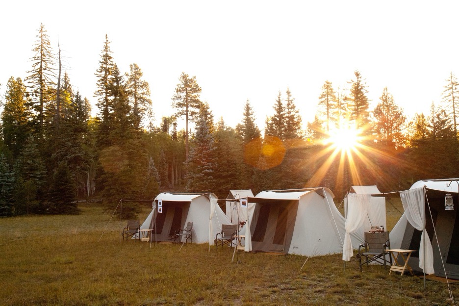 canvas tents sit in front of a forest with a sunrise behind them