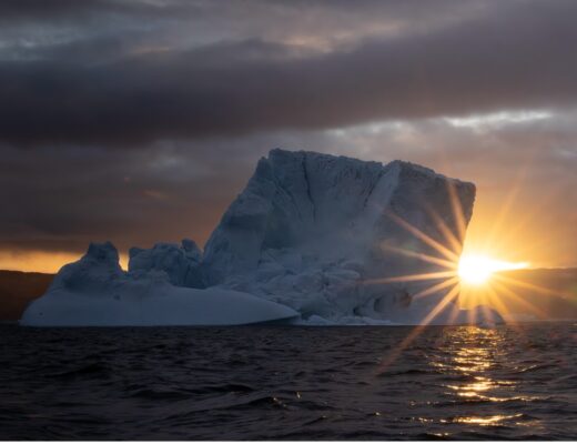 a sunburst appears on the horizon behind an iceberg in Greenland