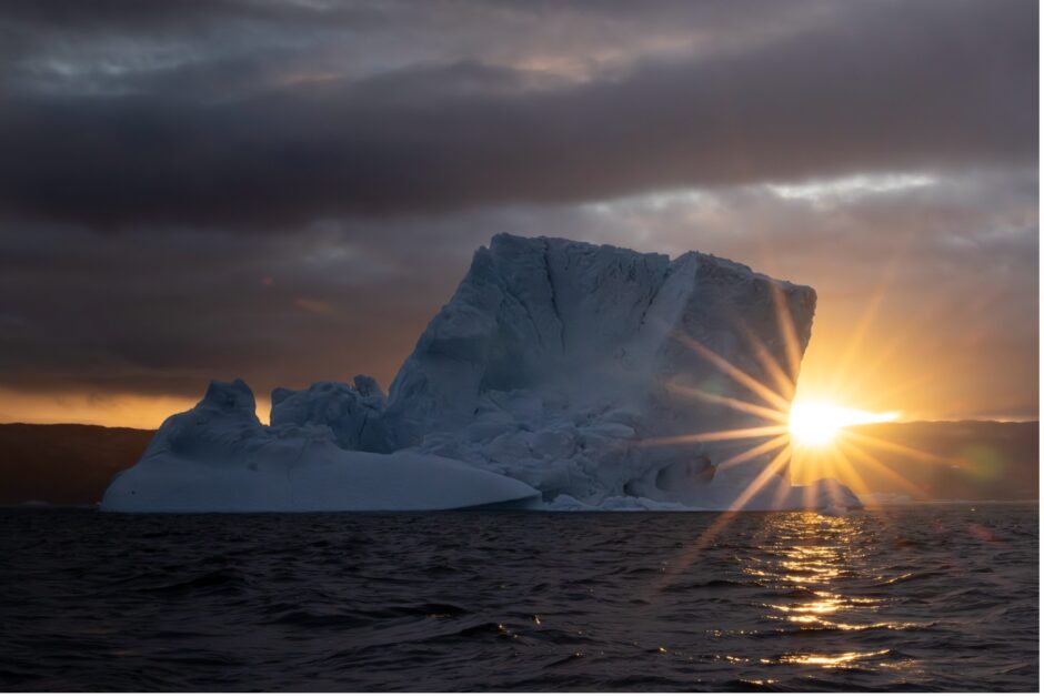 a sunburst appears on the horizon behind an iceberg in Greenland